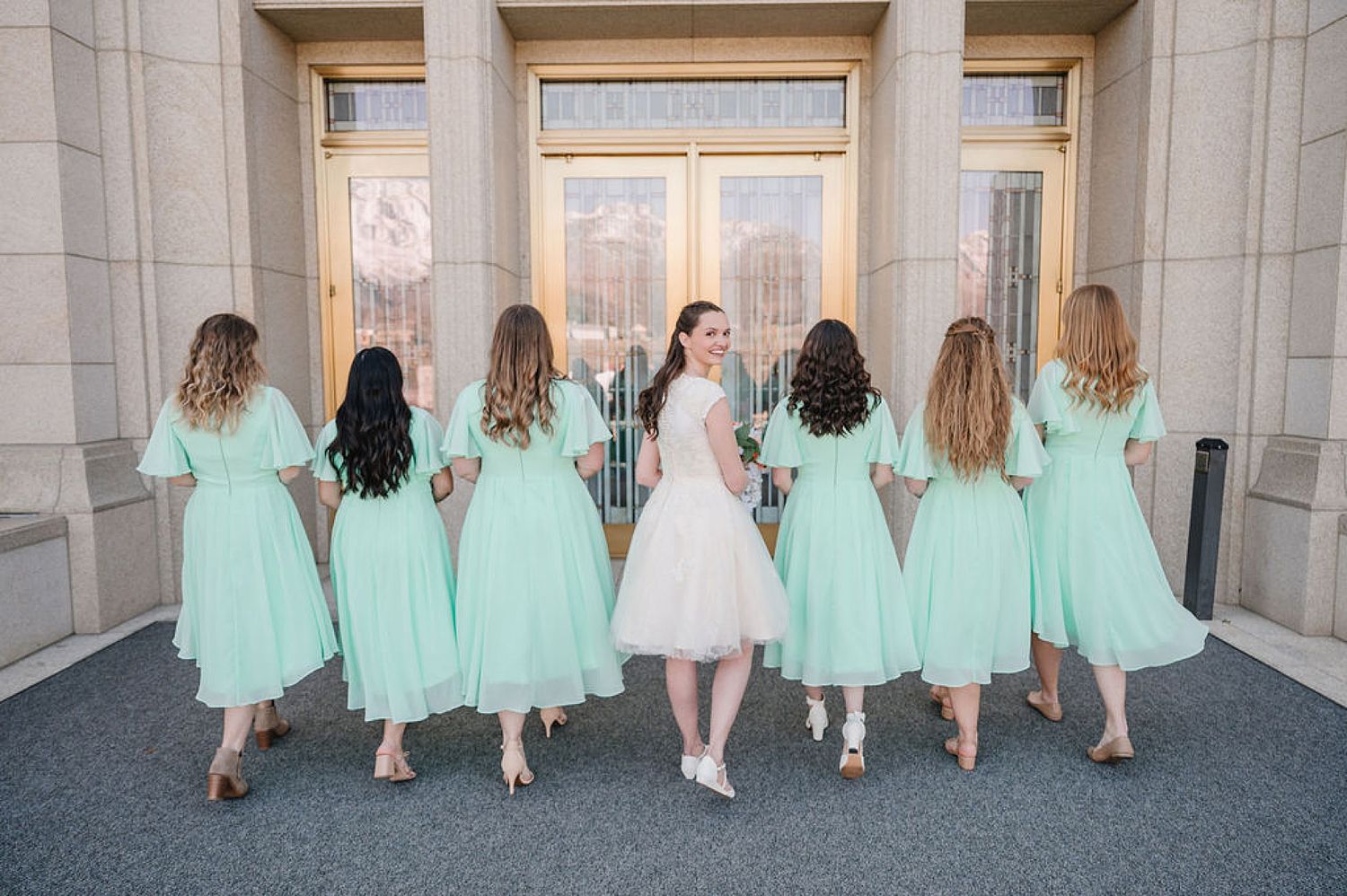 Bridesmaids in matching mint green dresses pose with backs turned at an elegant stone entrance.