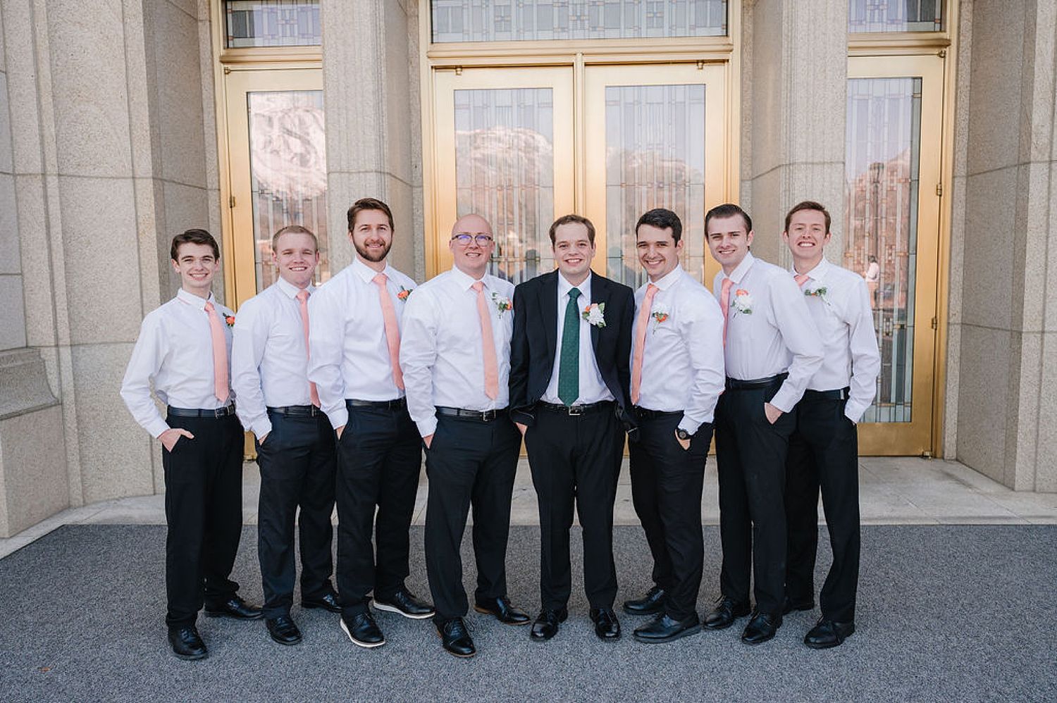 A group of formally dressed groomsmen in white shirts and black pants pose together outside elegant doors.