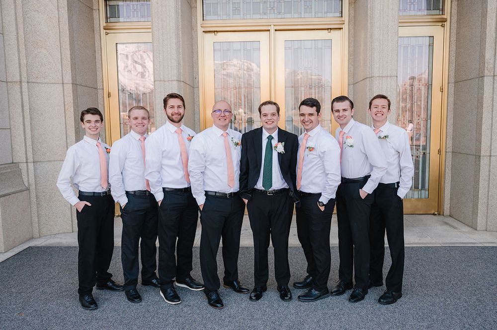 A group of formally dressed groomsmen in white shirts and black pants pose together outside elegant doors.