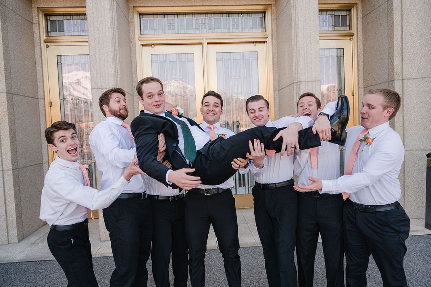 Groomsmen in white shirts and black pants playfully lift someone in formal attire at a building entrance.