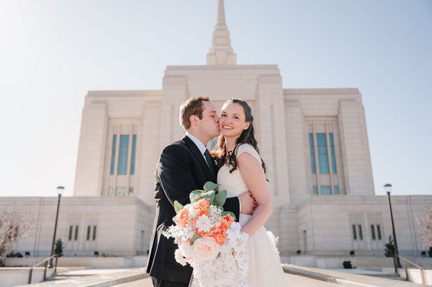 Newlywed couple shares a romantic moment outside a grand white Mormon temple with a coral and white bridal bouquet.