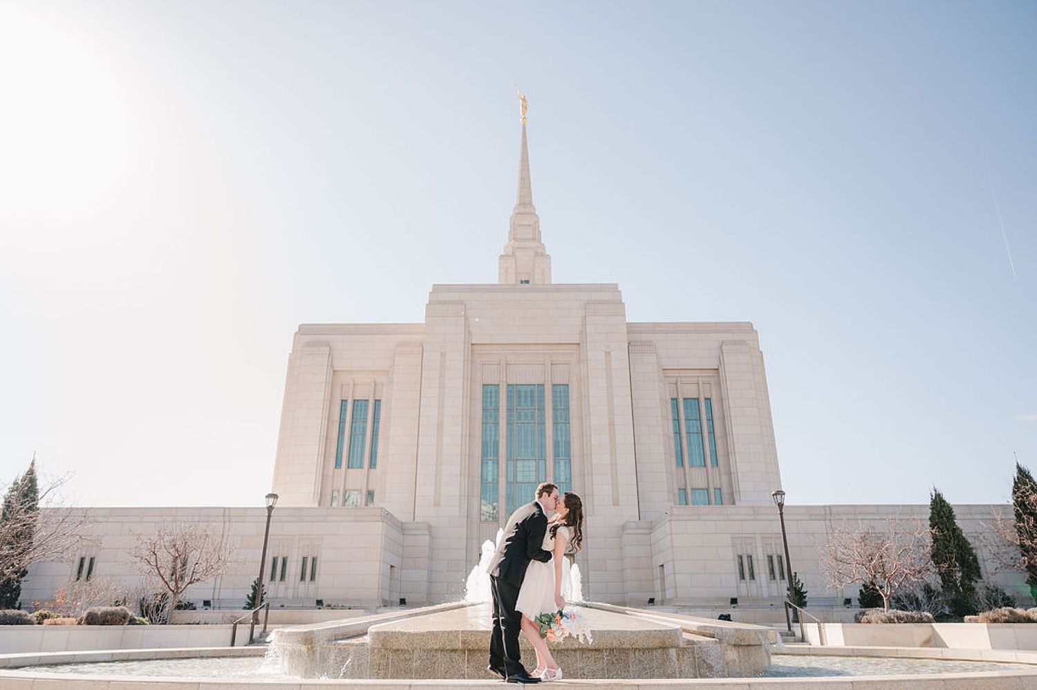 A couple embraces in front of the white marble Ogden Utah LDS Temple on a bright sunny day.
