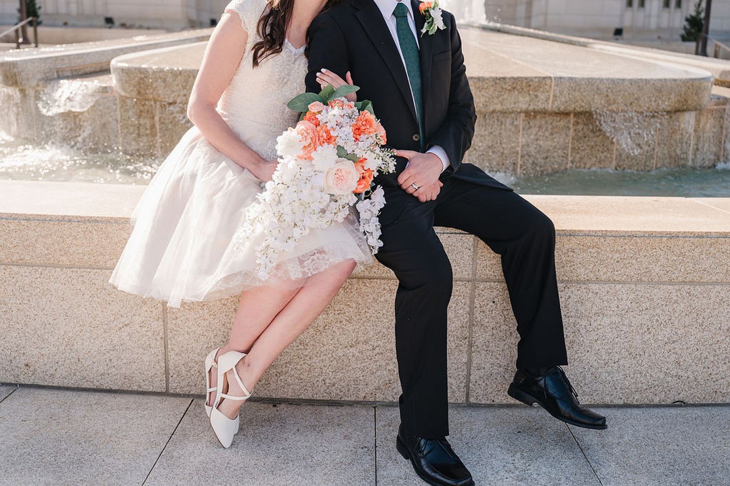 Couple sits on fountain edge showing wedding attire details with peach bouquet and formal shoes.