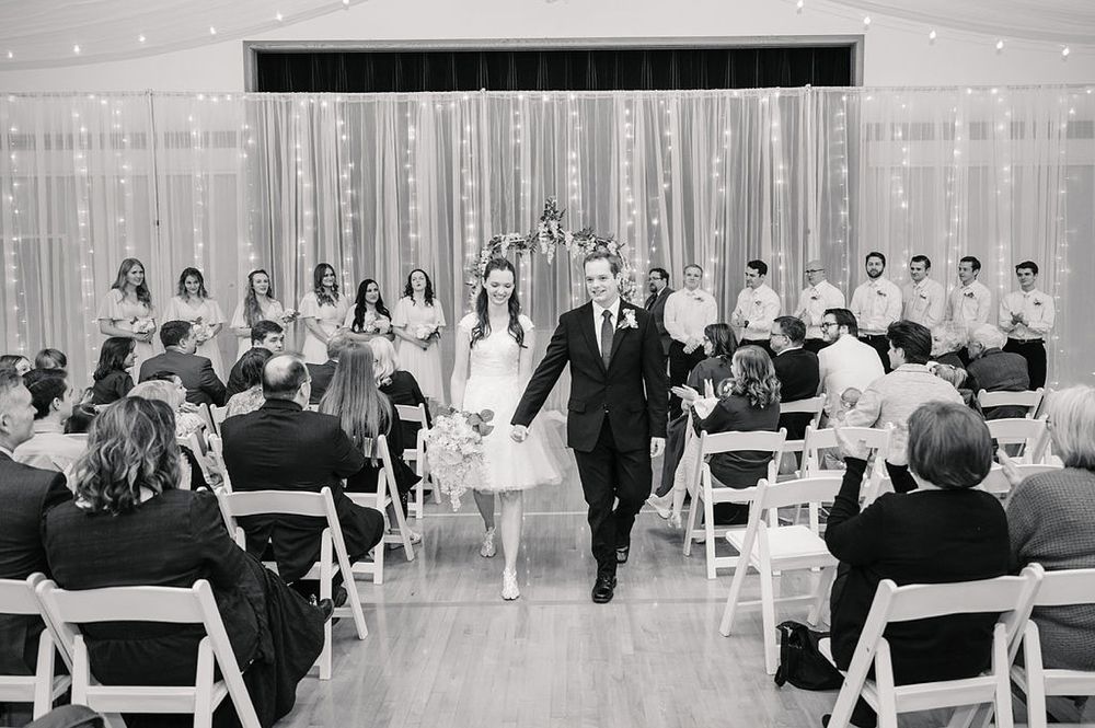 Black and white photo of a wedding ceremony in a hall with string lights and guests seated in white chairs.