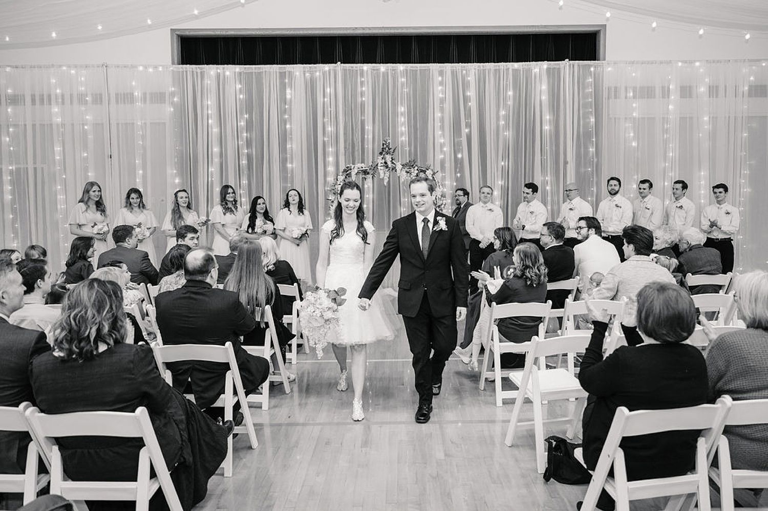 Black and white photo of a wedding ceremony in a hall with string lights and guests seated in white chairs.