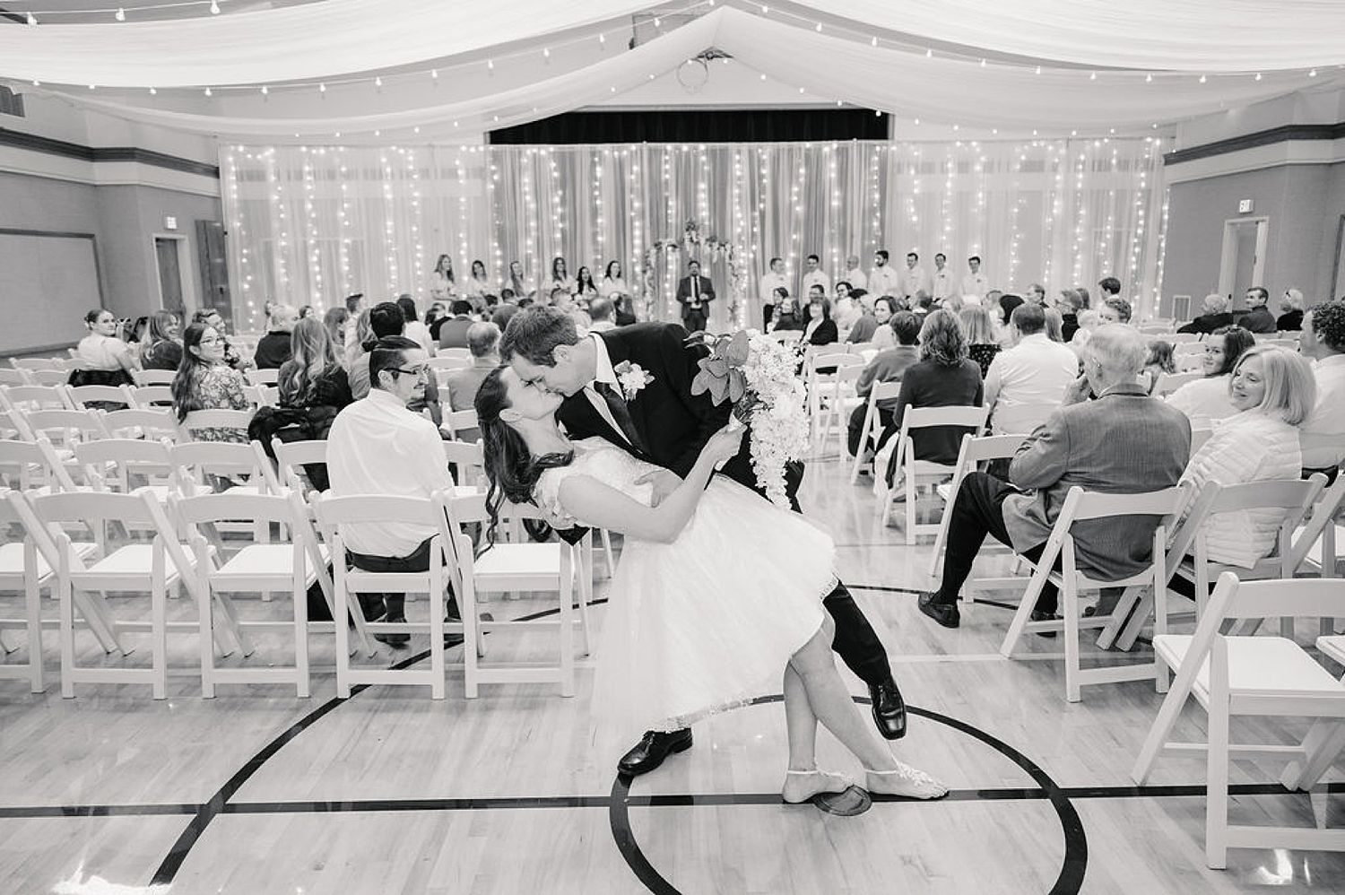 A romantic dip dance move by a couple during a wedding reception with guests seated in white chairs watching.