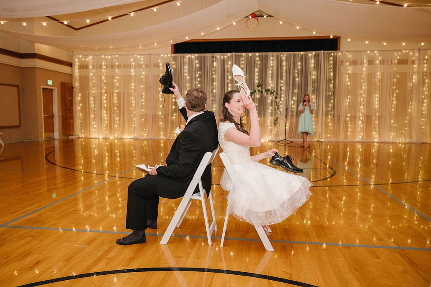 Two people sit back-to-back on chairs during a fun wedding game with glowing string lights in the background.