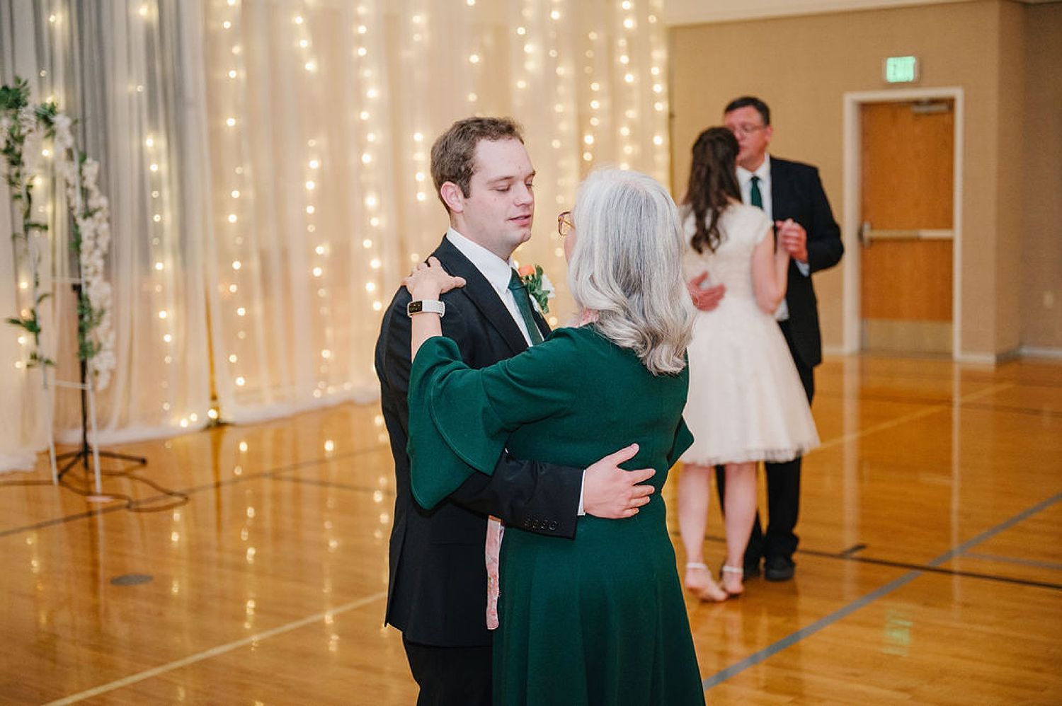 A couple dances together at an indoor wedding reception with string lights and wood flooring in the background.