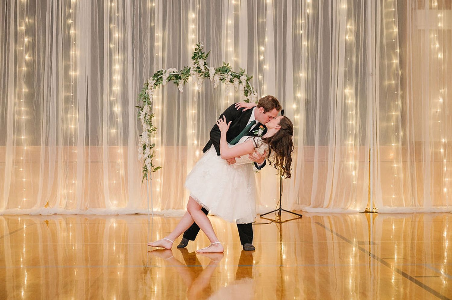 A romantic dance dip beneath a floral arch with twinkling lights creating a magical ballroom atmosphere.