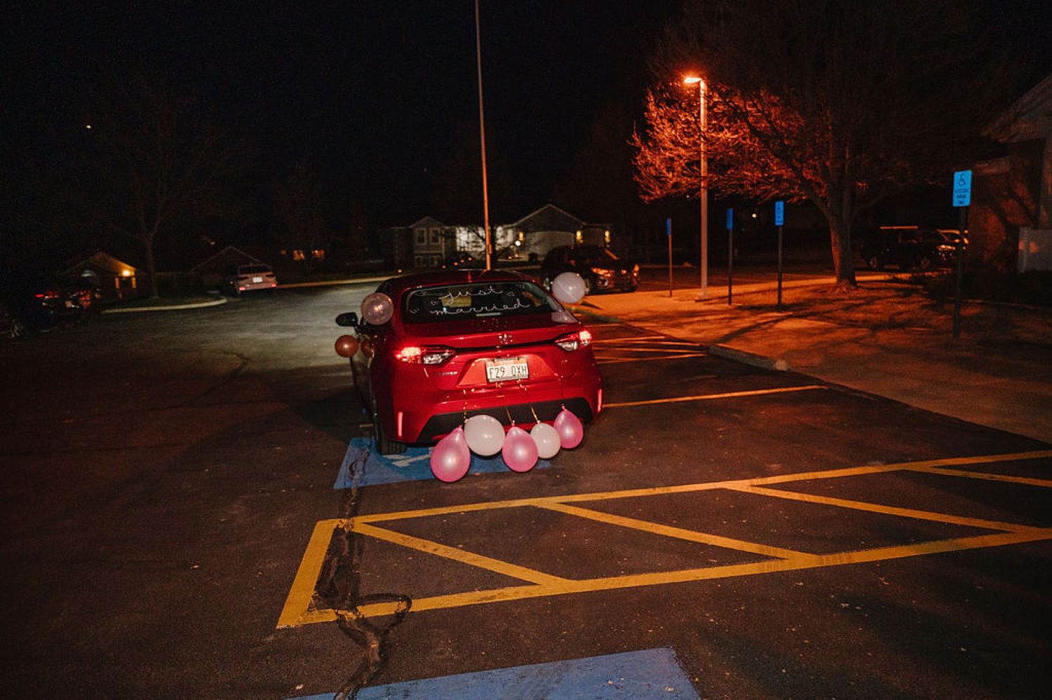 A red car parked in an illuminated parking lot at night with street lights glowing orange in the background.