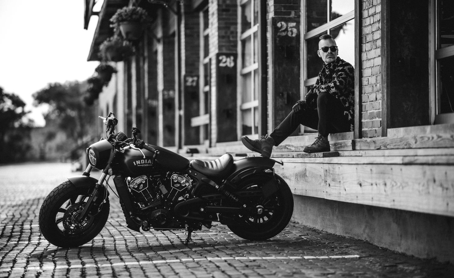 Black and white portrait with a custom motorcycle parked on cobblestone street in front of brick building facade.