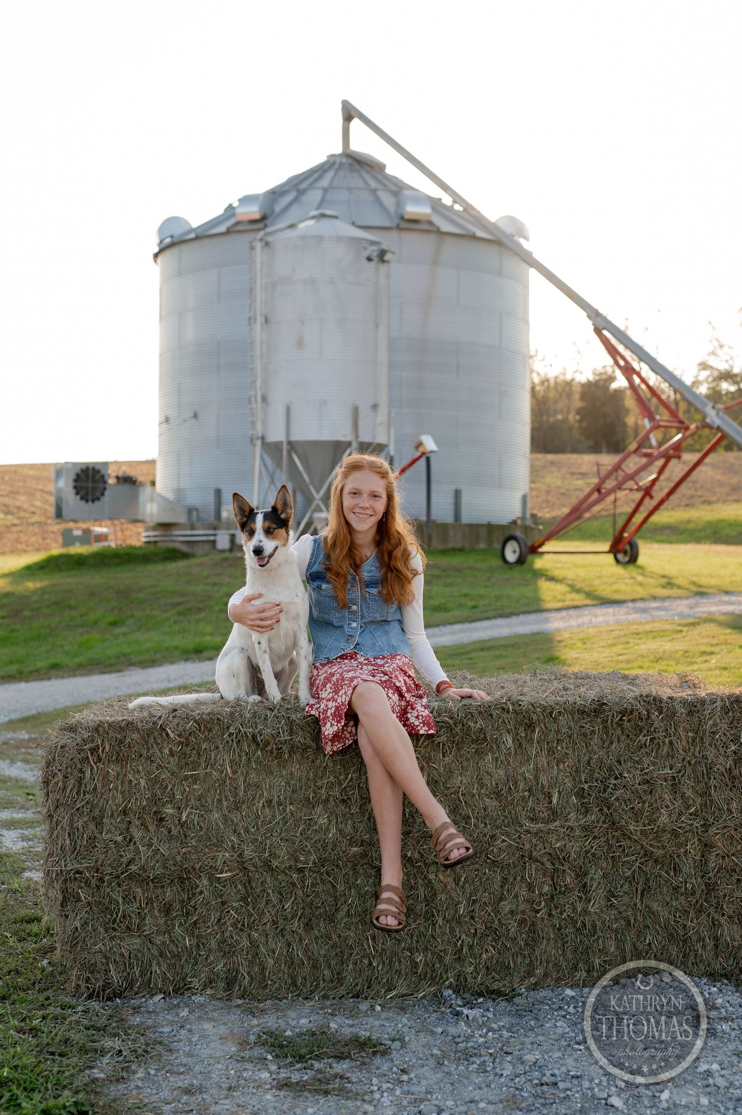 Person sitting with dog on hay bale near grain silo at sunset.