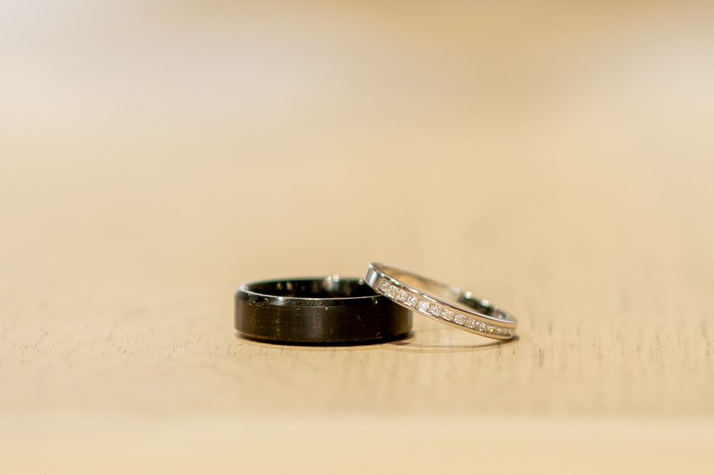 Two wedding rings, one black metal band and one diamond encrusted platinum band, displayed on a beige textured surface.
