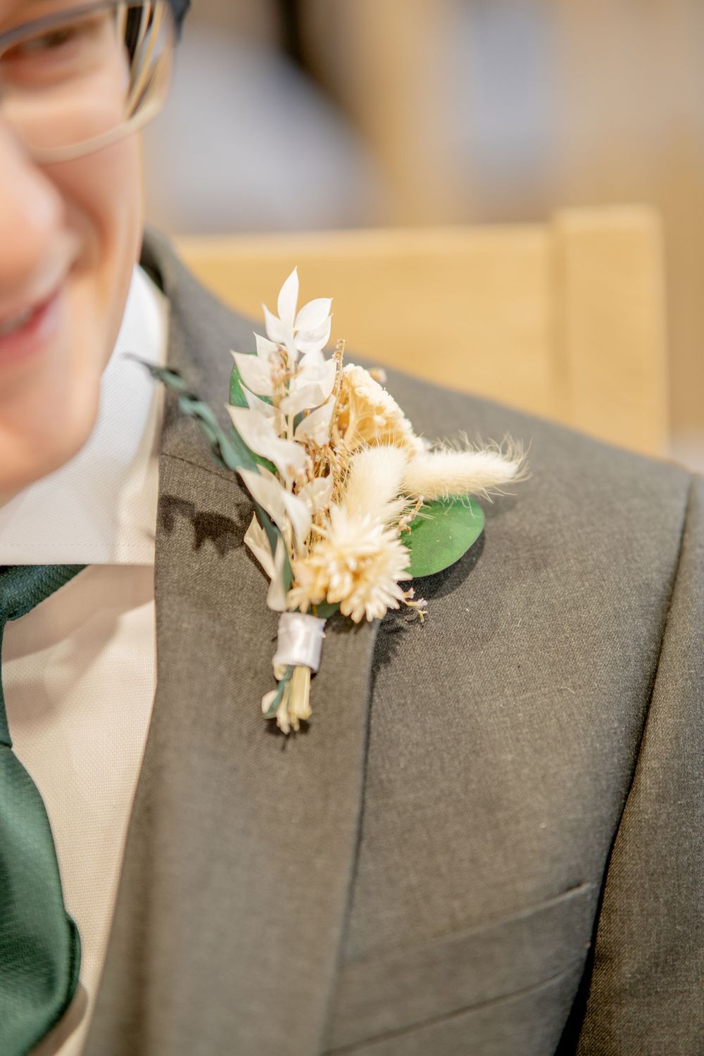 A delicate dried floral boutonniere with peach and white blooms pinned to a grey suit lapel.