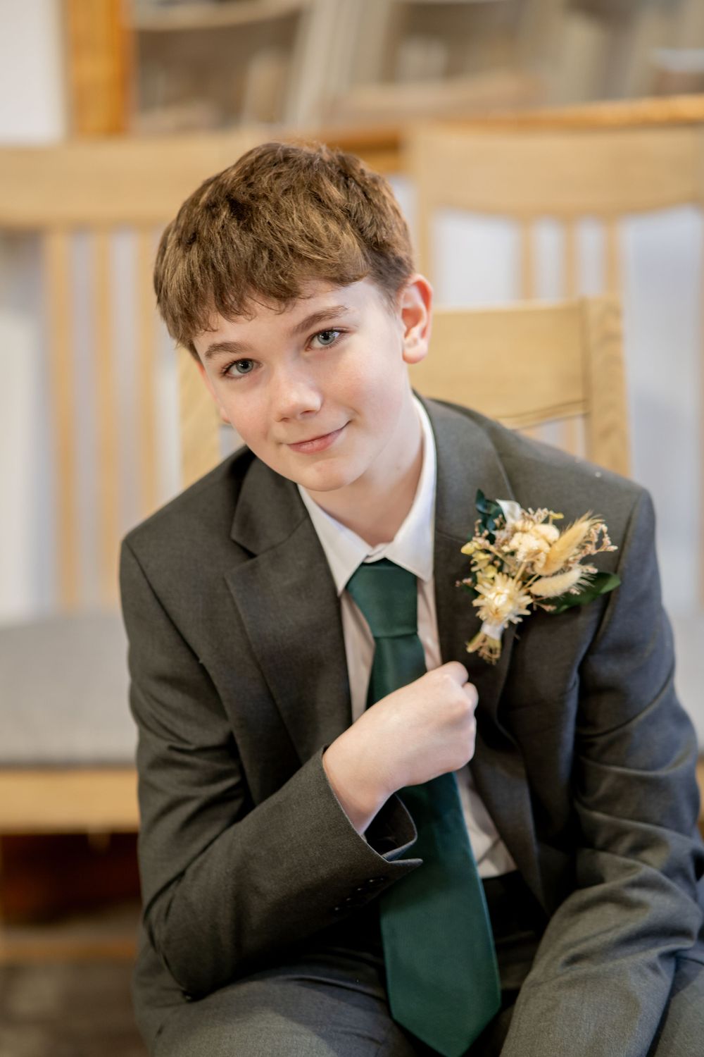 Young guest in formal dark suit with emerald green tie and floral boutonniere sits in wooden chair.