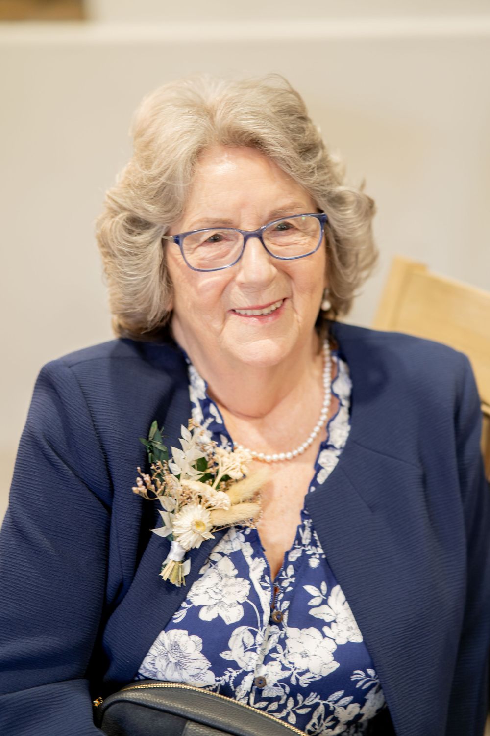 Elegant woman wearing navy blazer and floral dress with pearl necklace and corsage at formal event.