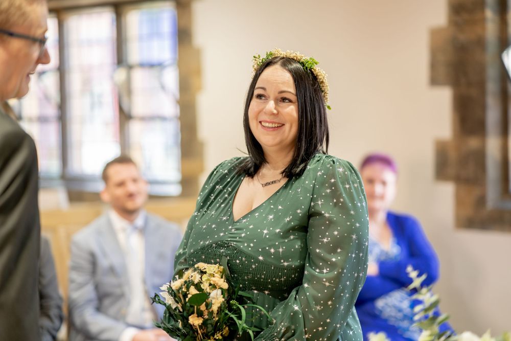 A bride wearing a sparkly green dress and floral crown smiles during an indoor ceremony.