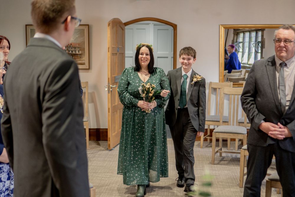 Wedding party moment shows bride in emerald green polka dot dress walking down aisle at intimate indoor ceremony.