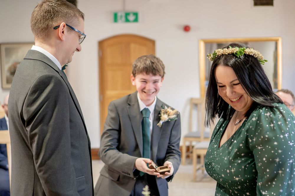A wedding ceremony shows the exchange of rings between a couple wearing green and grey attire in an indoor venue.