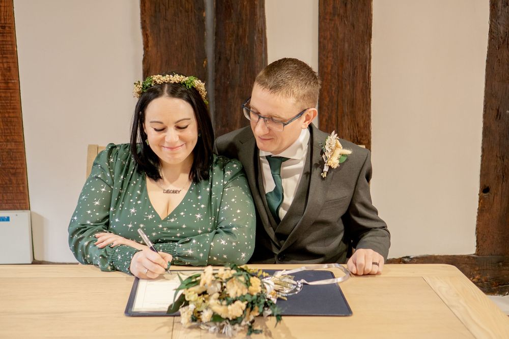 A couple in green and grey attire signs documents during their wedding ceremony at a wooden table.