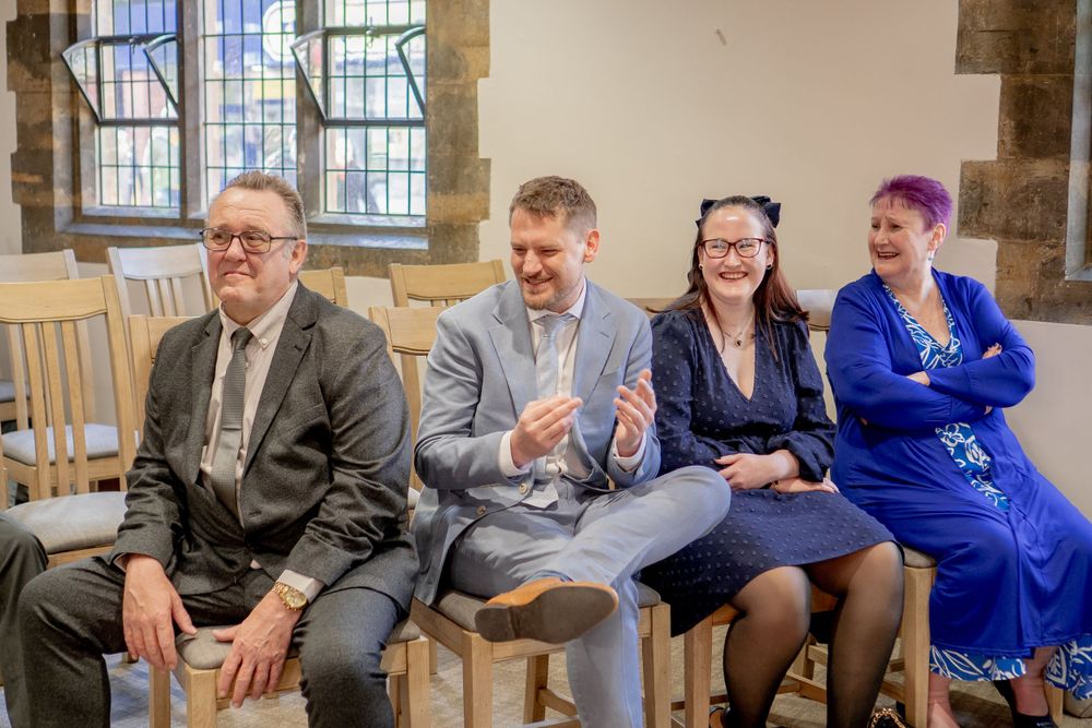 Four people sit together on wooden chairs sharing a lighthearted moment in what appears to be a historic building.
