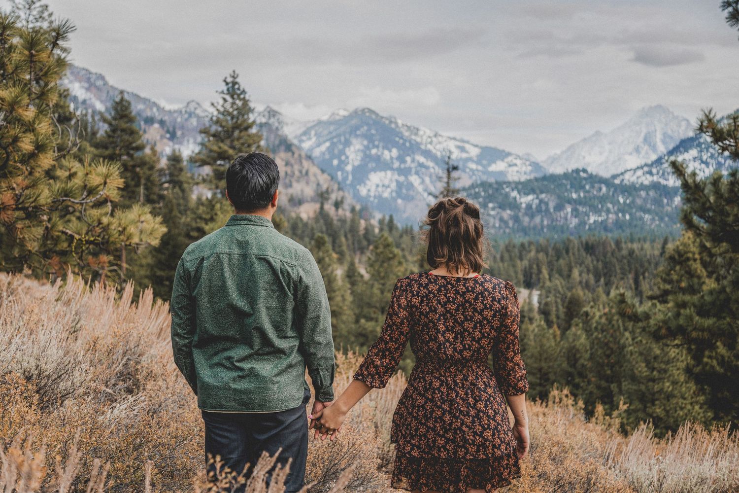 Two people admire a scenic mountain landscape with snow-capped peaks and evergreen forest from a hiking trail.