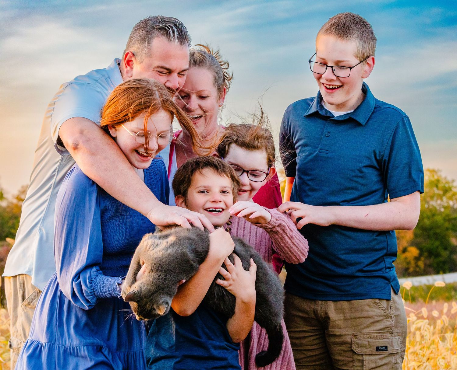 A family shares a joyful group hug together outdoors during golden hour with a blue sky background.