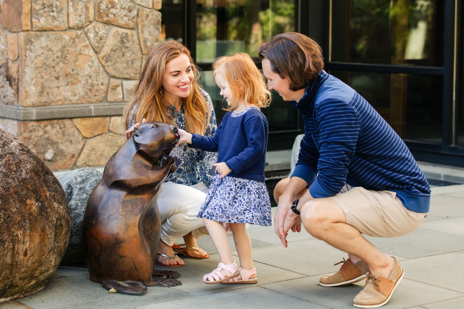 Family crouches down on stone patio to pet and interact with friendly brown dog in outdoor setting.