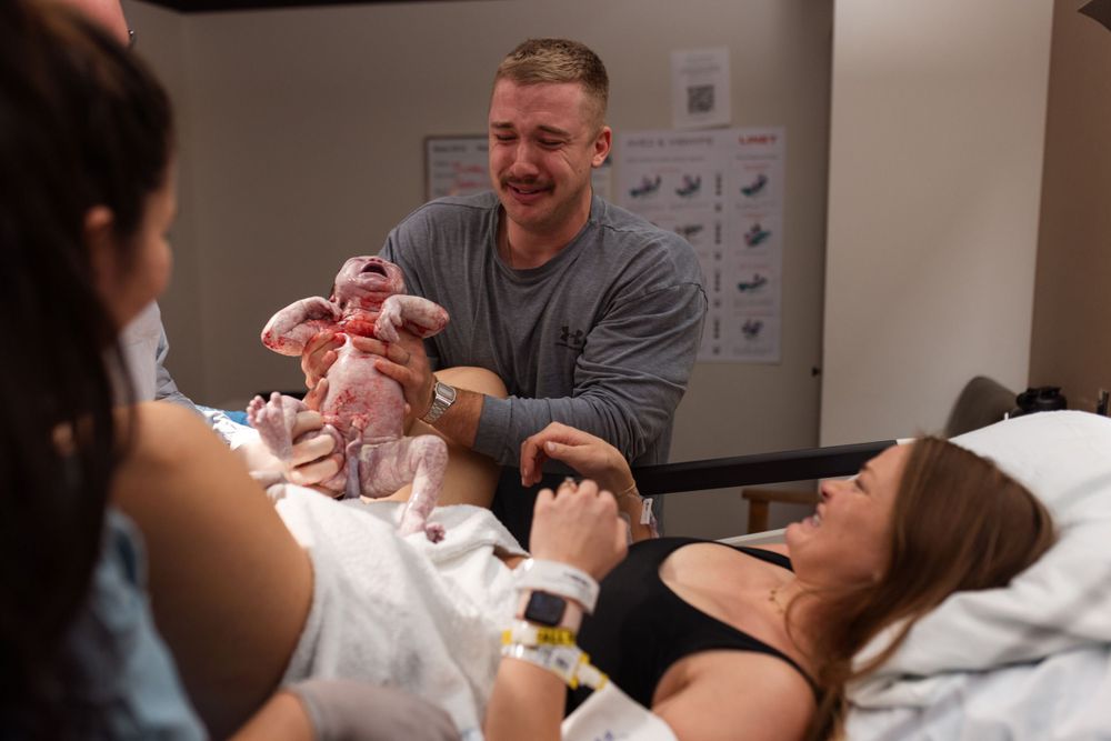 A newborn baby being held in a hospital room while medical monitors display vital signs in the background.