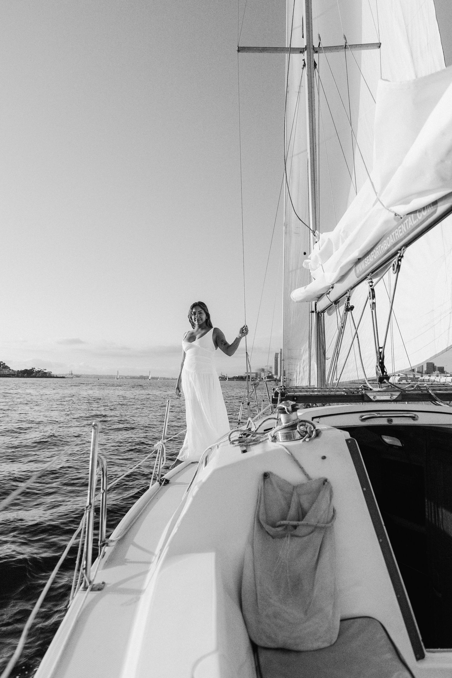 Black and white photograph of a figure in a white dress standing on a sailing yacht deck against a coastal backdrop.