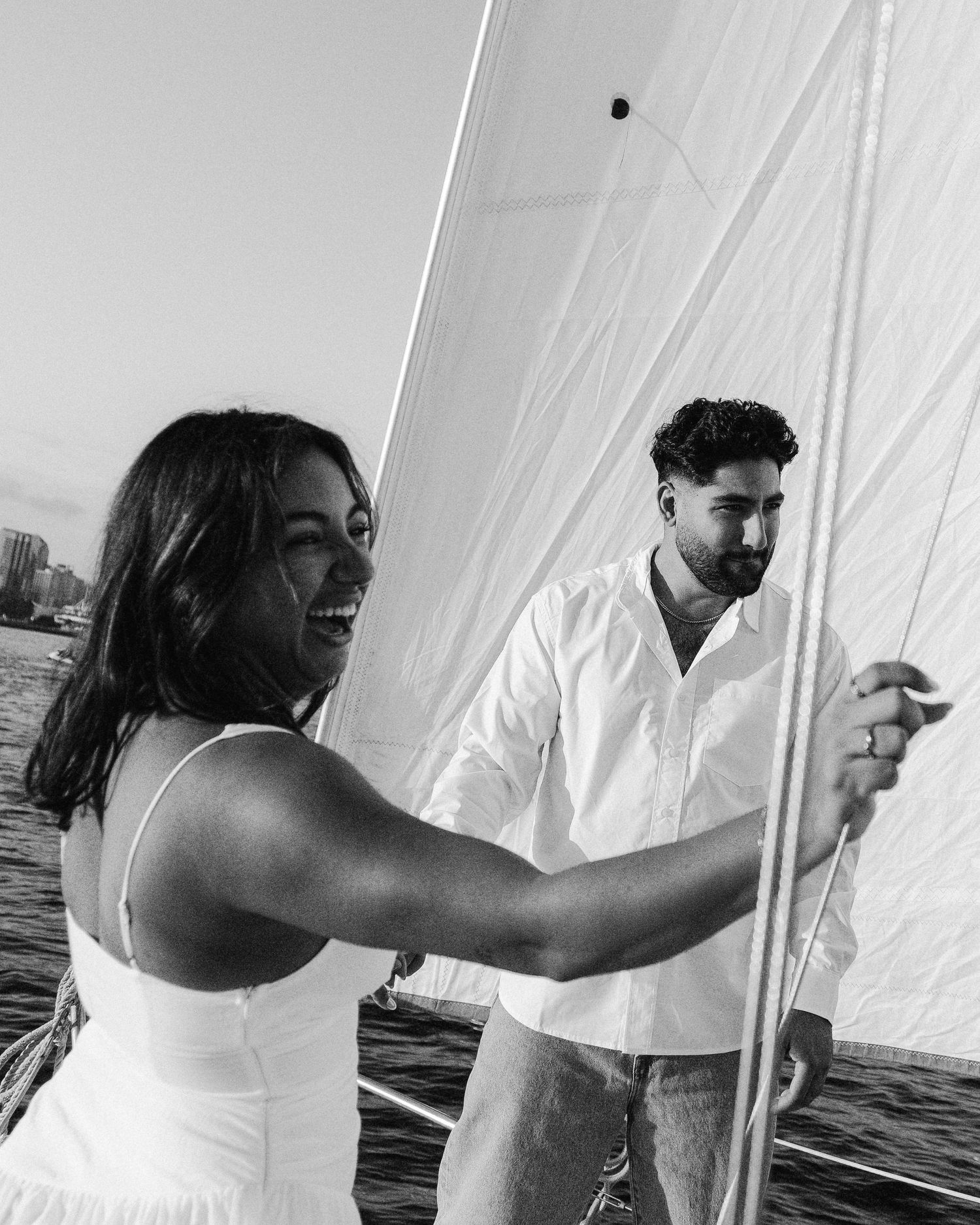 A couple in white outfits stand near sailboat rigging on a boat in black and white photography.