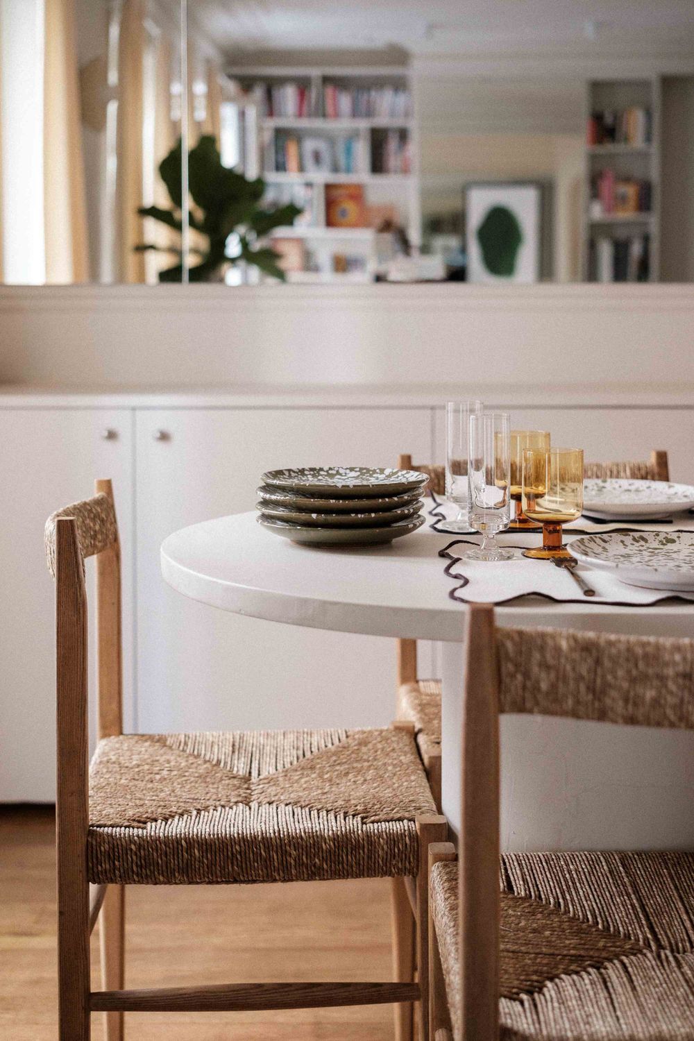 Close-up of white dining table with woven chairs and stacked plates against built-in bookshelf.