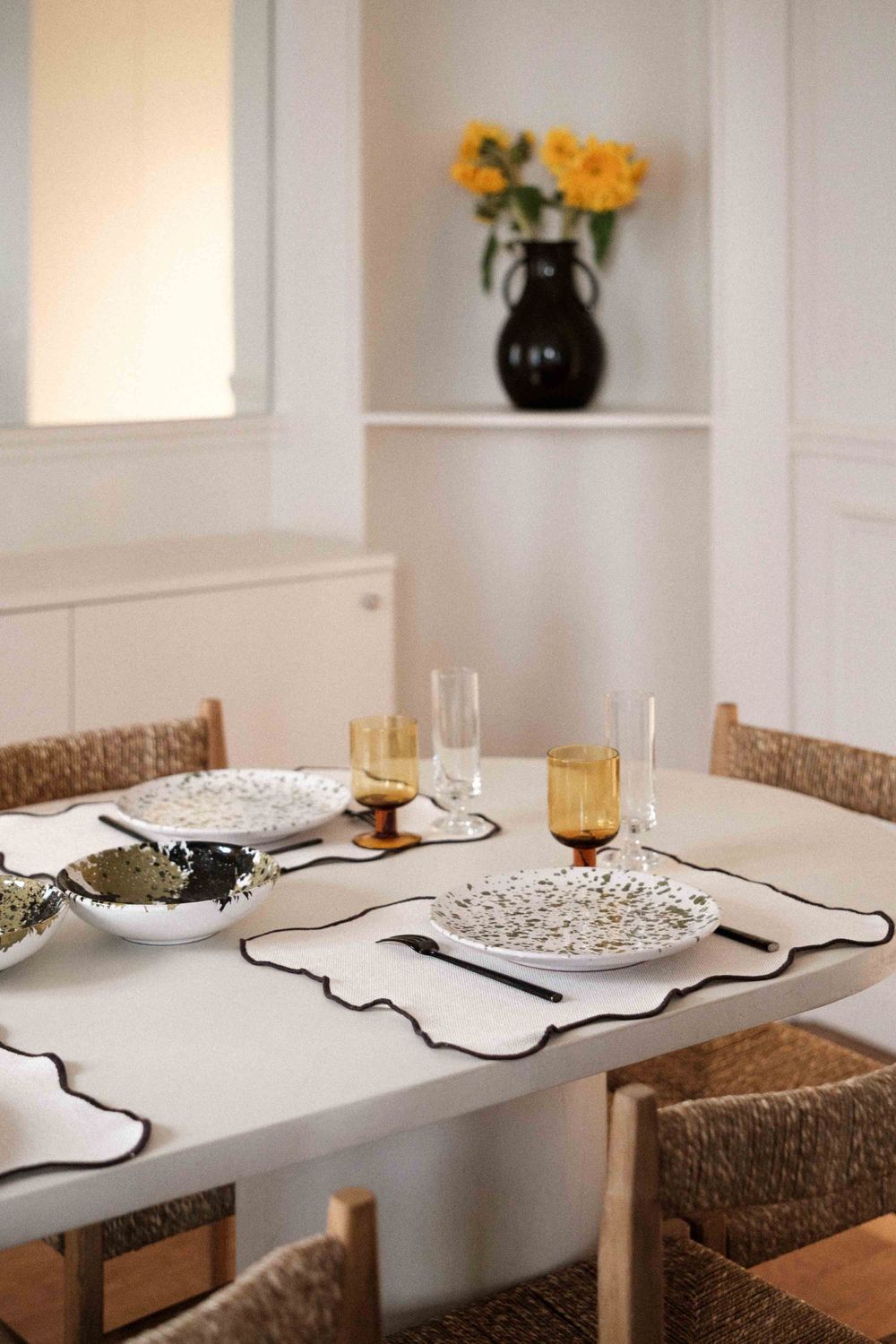 Elegant dining table setting with white tablecloth, woven chairs, and yellow flowers in black vase.