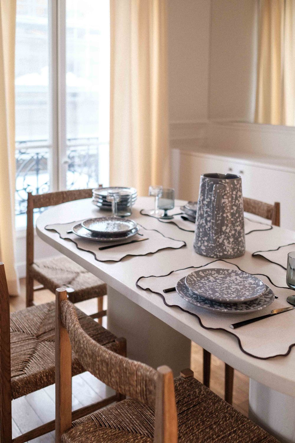 Bright dining room with woven chairs and speckled plates set on white table.