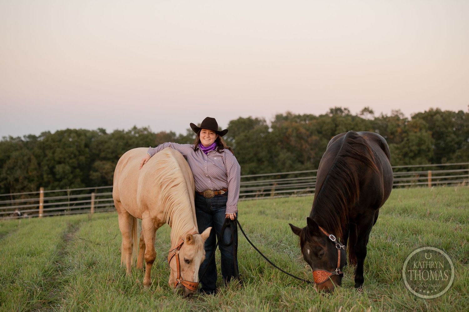 Two horses eating grass beside their handler in a fenced pasture during golden hour.