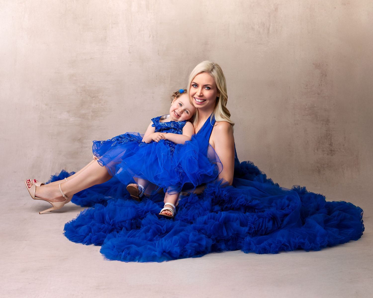 Mother and daughter sitting in matching dresses, daughter sits on mothers lap both smiling sweetly, in studio setting.