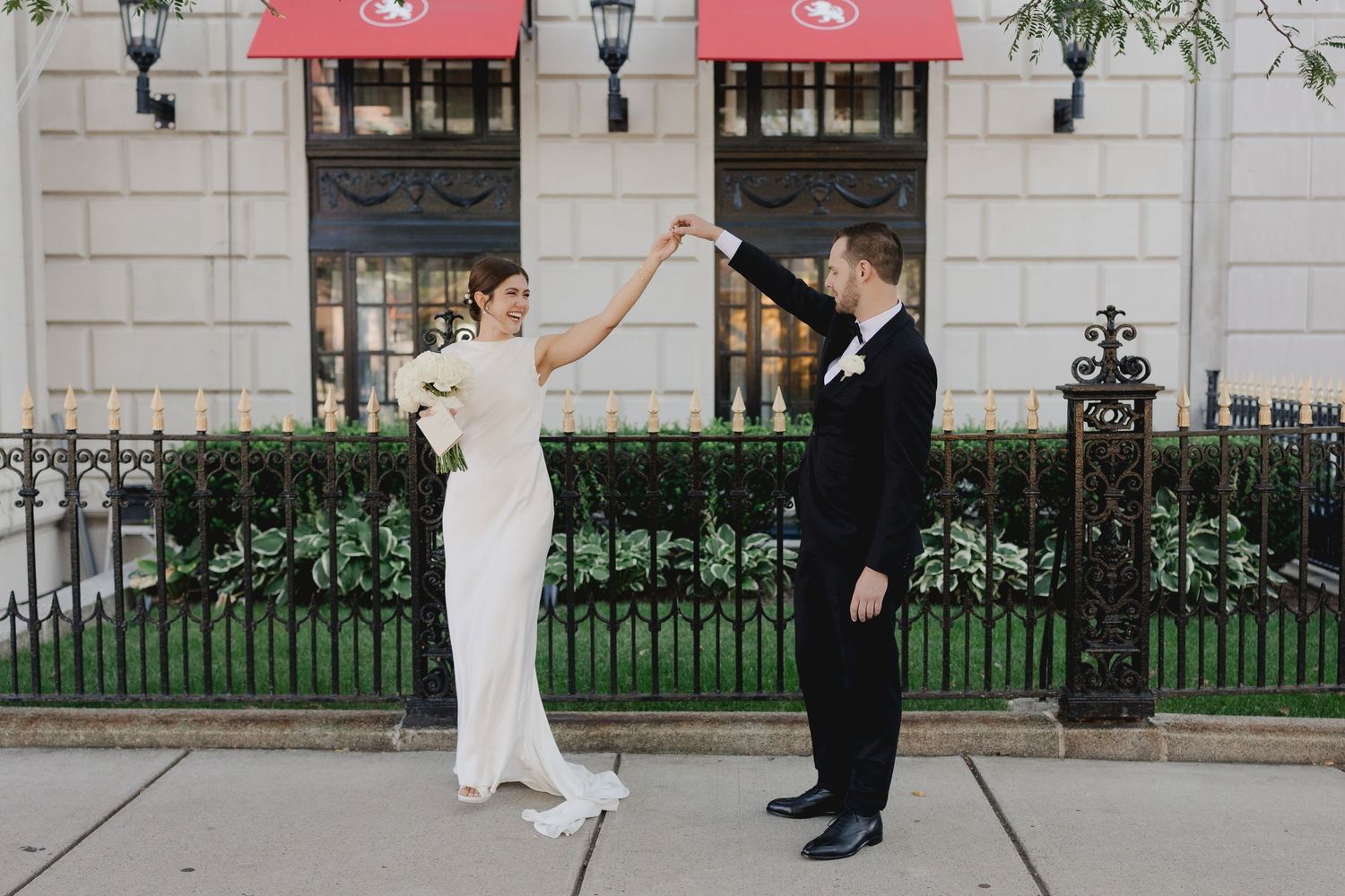 Bride and groom dancing on sidewalk outside elegant building with red awnings, groom spinning bride in wedding dress.