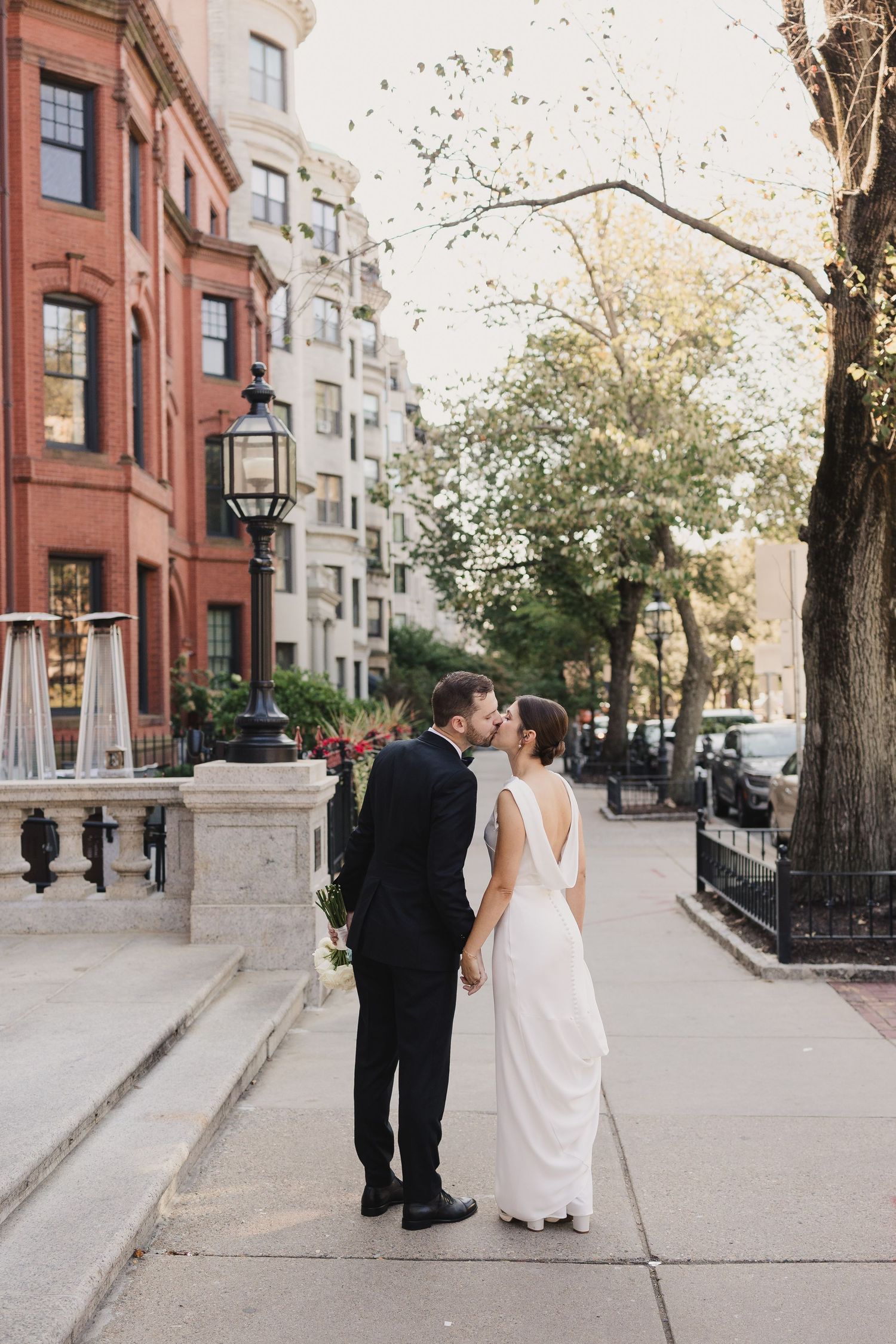 Newlyweds sharing a romantic kiss on a tree-lined urban sidewalk, bride in elegant white gown.
