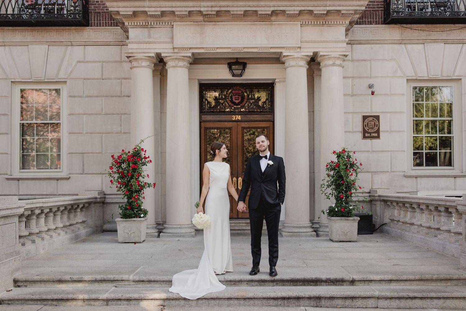 Bride and groom hold hands on elegant stone steps of a classic building, surrounded by red roses.