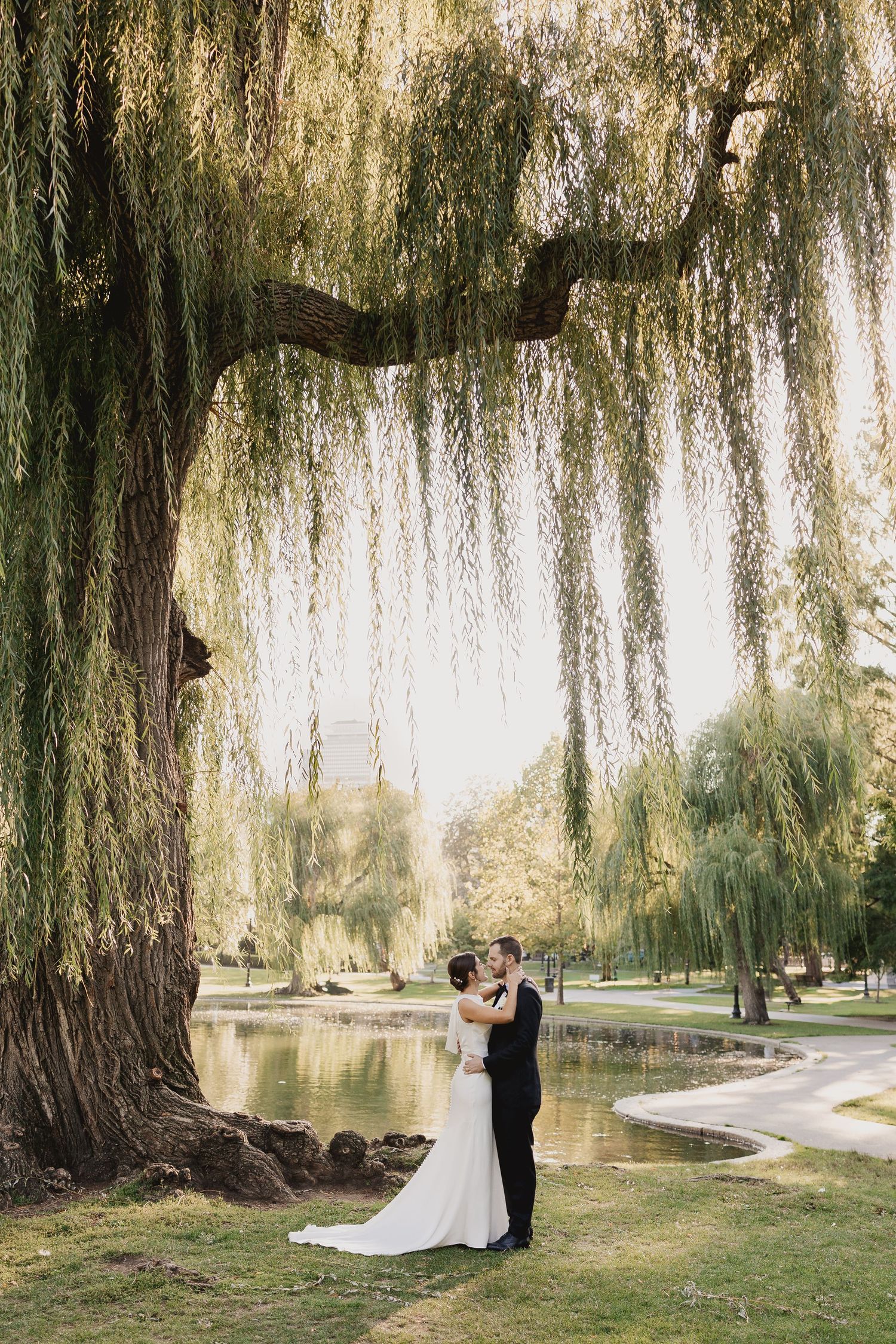 Bride and groom share a romantic kiss under a weeping willow tree by a serene pond in a lush green park.