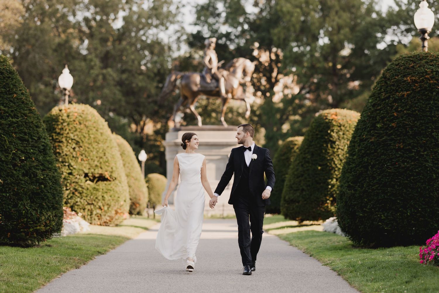 Bride and groom walk hand-in-hand through elegant garden path with sculpted hedges and equestrian statue backdrop.