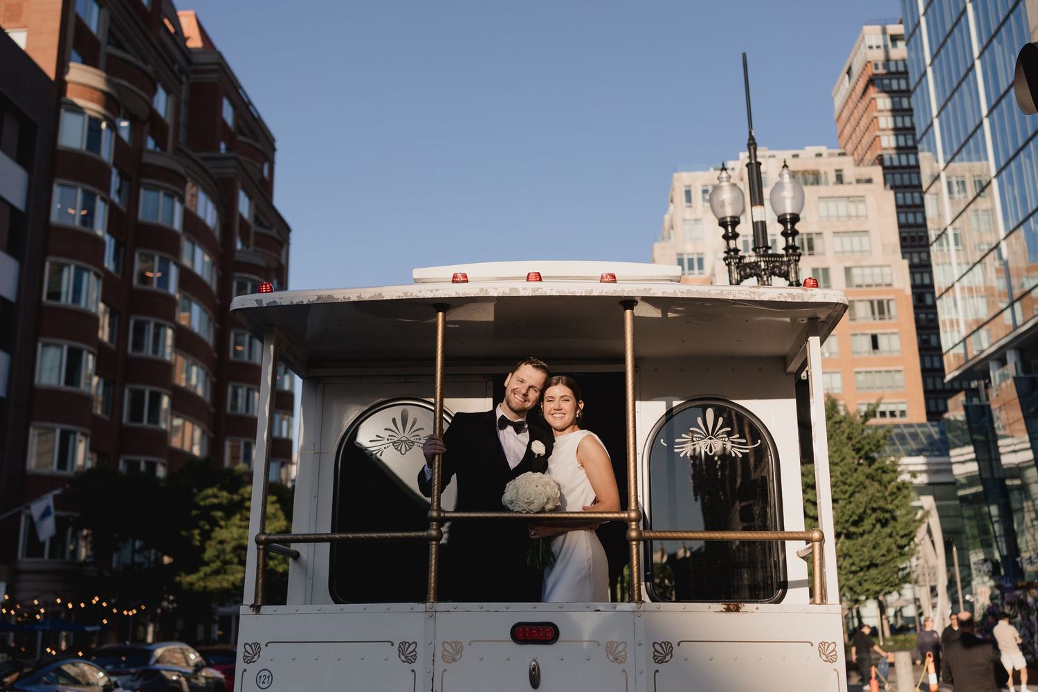 Bride and groom smile together aboard a white trolley with chandelier in an urban cityscape.