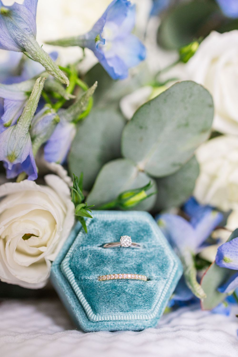 Diamond engagement ring and wedding band nestled in teal velvet ring box surrounded by white roses and blue flowers.