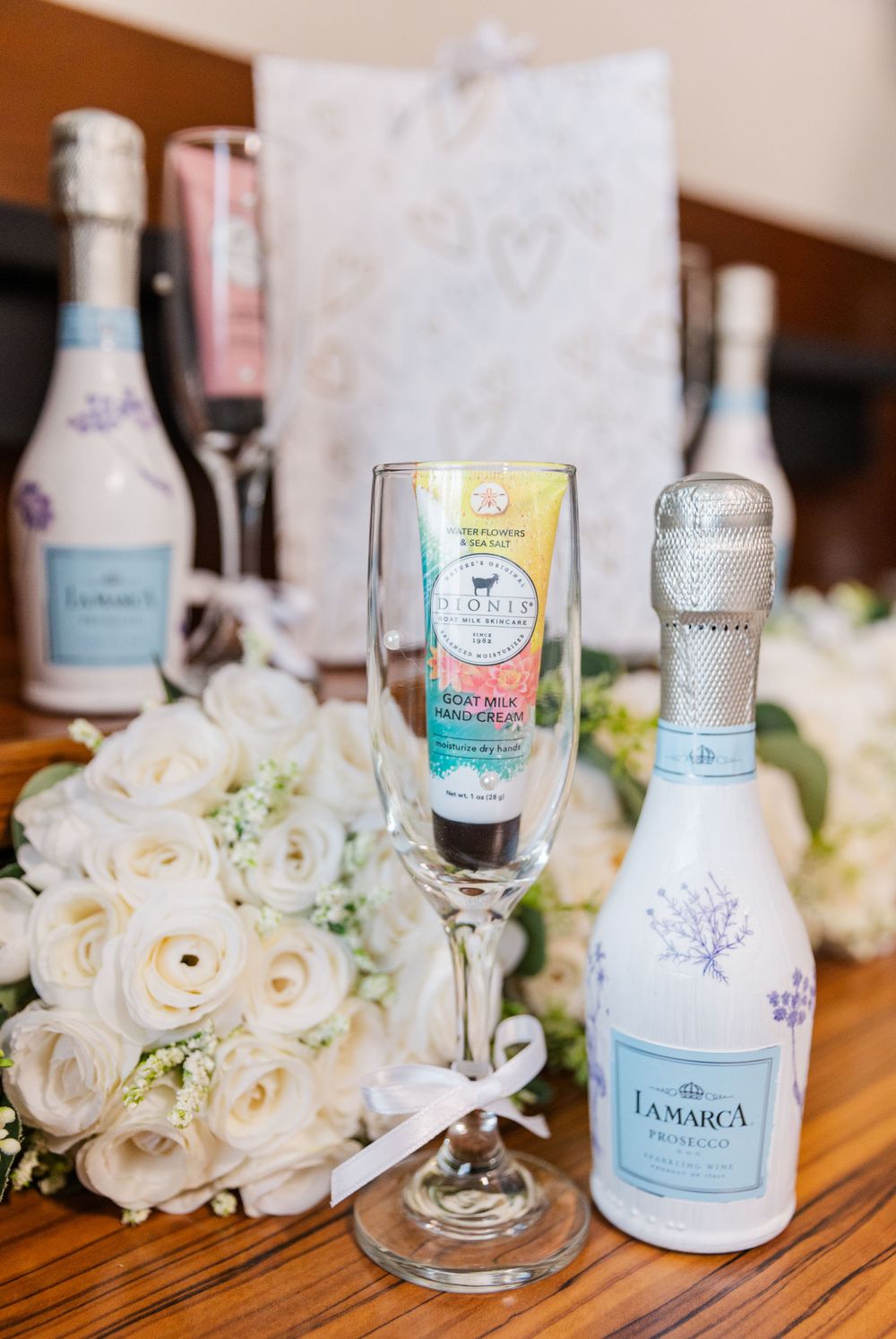 Wedding reception details with champagne bottle, floral arrangements and decorative glassware on wooden table.