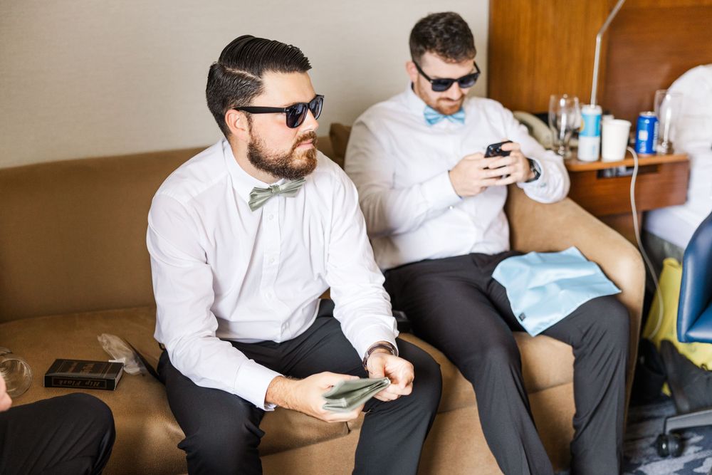 Two groomsmen in white shirts and sunglasses relaxing on a couch while looking at their phones before the wedding.