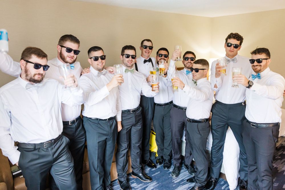 Group of groomsmen in matching white shirts and sunglasses raising drinks in celebration.