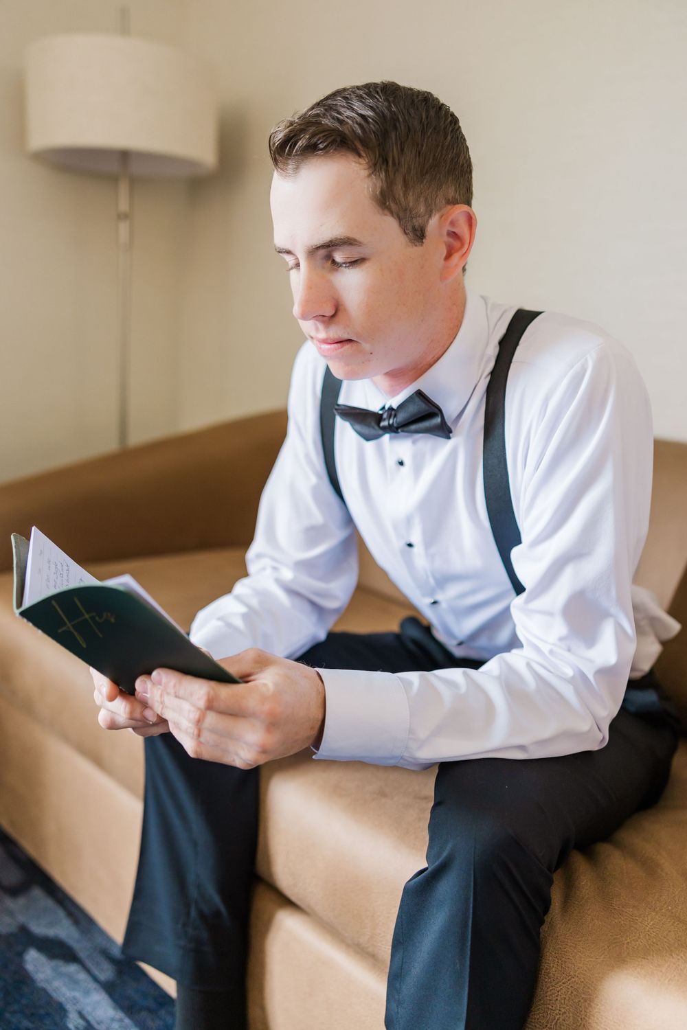 Groom in formal attire with bow tie and suspenders reading from a small book while seated on couch.