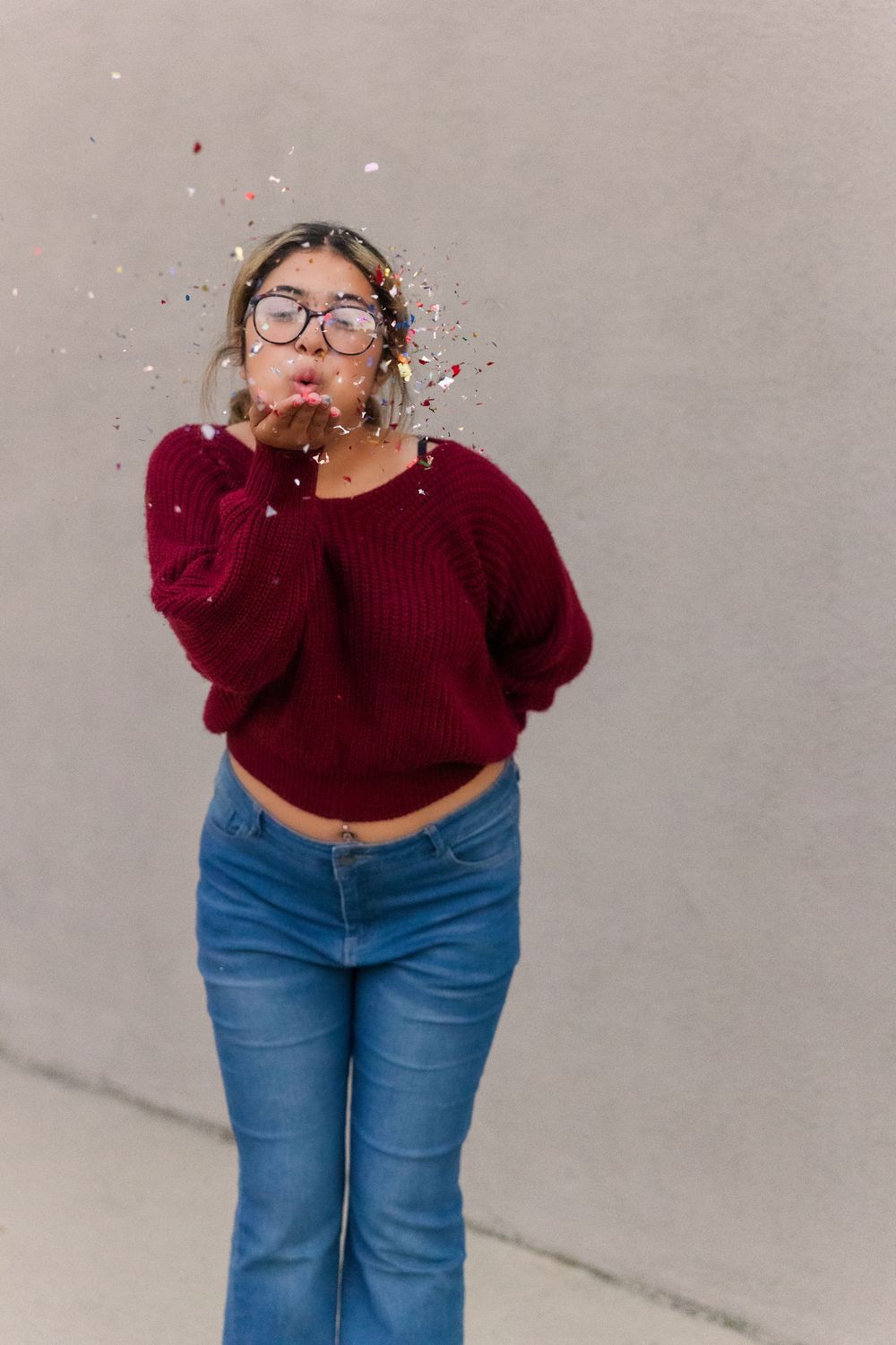 Someone in a burgundy sweater and jeans blows confetti while posing against a gray wall.