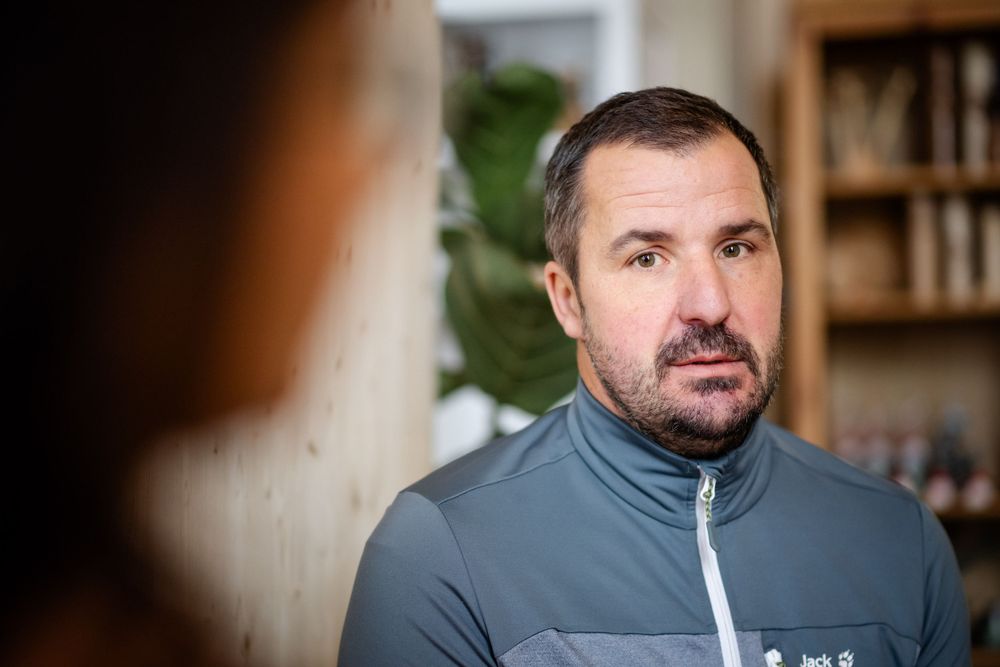 Professional wearing a gray athletic jacket in an office setting with bookshelves in the background.