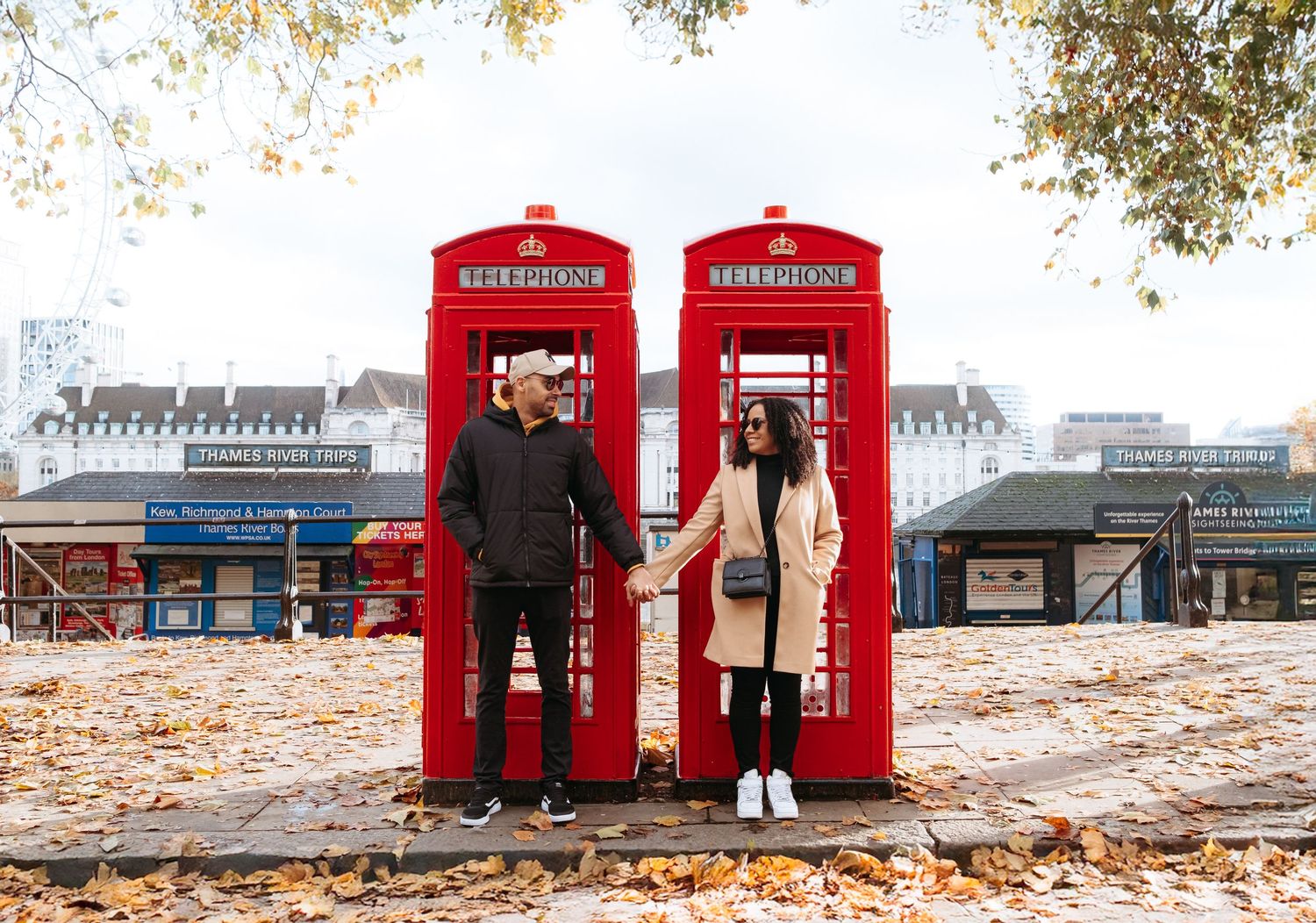 Two people pose in iconic red London phone booths surrounded by autumn leaves.